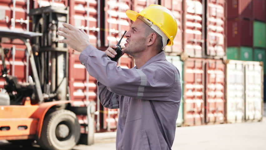 Two male workers in safety helmets examine and discuss a shipping container during operations at a busy logistics terminal.