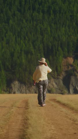 Person walks down a dirt path through a field. The person wears a hat and carries a bag. They appear focused, moving steadily forward. Lush green forest lines the background. Footage suits travel