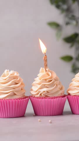 Close-up of cupcake in a bright pink liner with tall buttercream frosting swirl and a lit gold candle, cupcake in a pink cupcake liner with vanilla buttercream on beige background