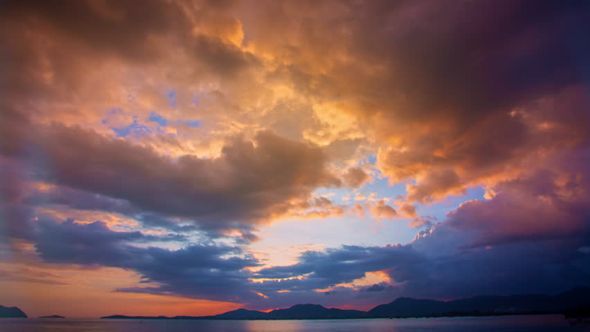 Clouds drift gently above the mountains and sea as the sun slowly disappears beyond the horizon. Golden sunlight paints the sky with beautiful colors while the ocean sparkles with shimmering reflections.