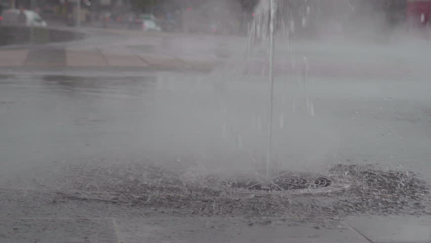Close-up of water jet spraying upward from ground fountain, mist and ripples forming on wet pavement in urban public space