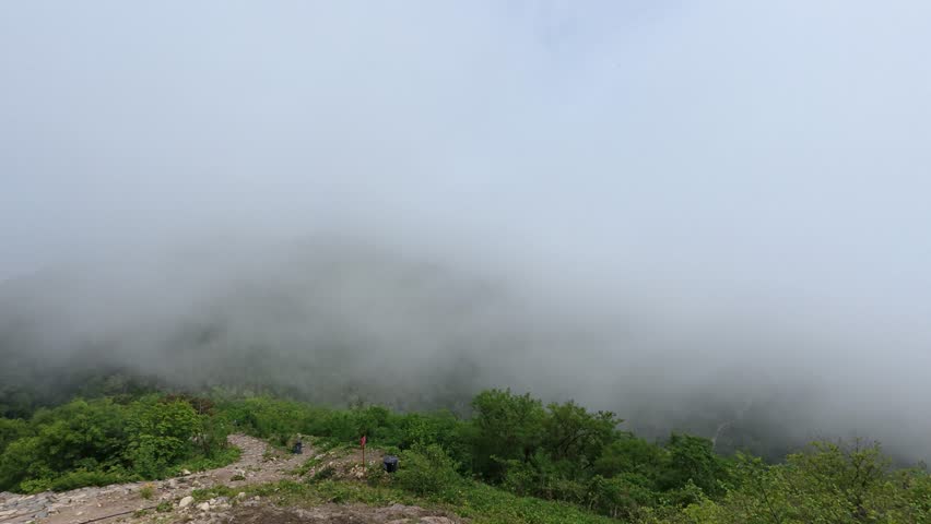 This landscape shows a dirt hiking trail atop a lush green hill, with thick fog obscuring the distant mountain peaks under an overcast pale sky.