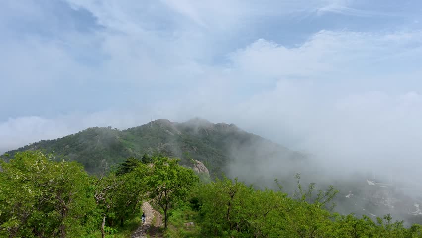 This scenic outdoor view features a winding hiking trail through lush green woodland, with misty fog rolling over the forested mountain peaks under a partly cloudy blue sky.