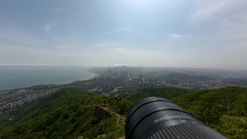 A high-angle panoramic shot taken from a mountain summit, showcasing a coastal city, adjacent beach, forested hills, and a clear daytime sky with a viewing telescope in the foreground.