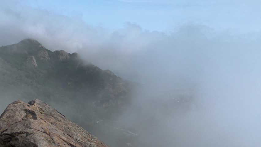 This landscape shows rugged mountain peaks partially shrouded in thick drifting mist and clouds, with a clear patch of blue sky visible above.