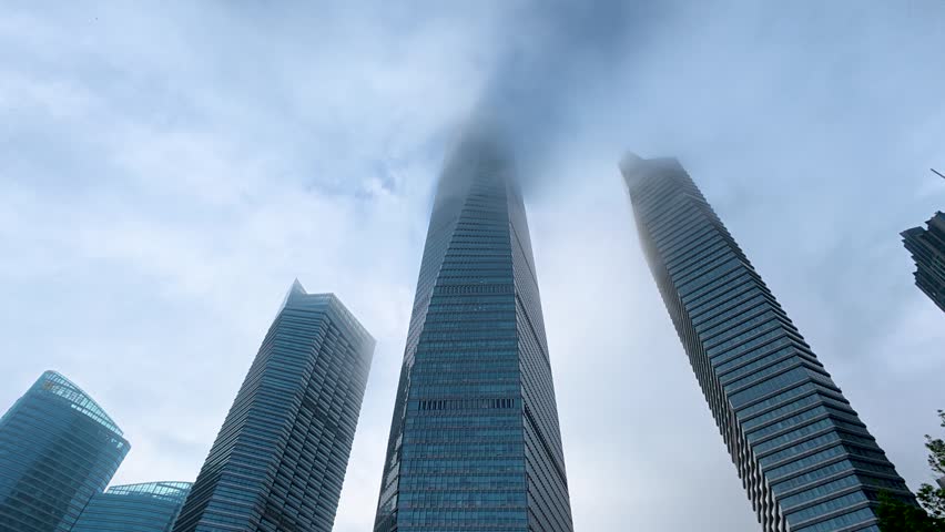 Low-angle shot of modern high-rise buildings shrouded in fog and haze over a cloudy urban sky.