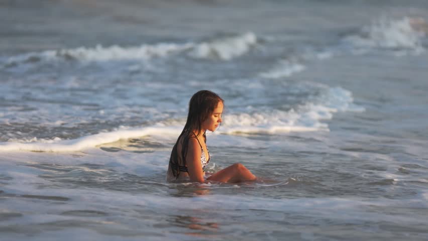 Girl with wet hair in a leopard swimsuit splashes water while sitting in the sea