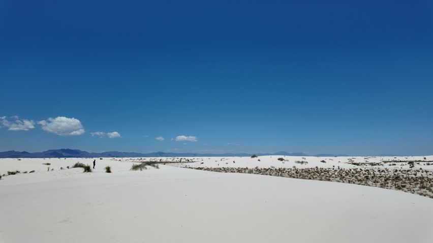 Vast Dunes at White Sands National Park Under Blue Sky - Vast white sand dunes stretch towards distant mountains under a clear blue sky at White Sands National Park, New Mexico. Sparse desert vegetation dots the bright, sunlit landscape.