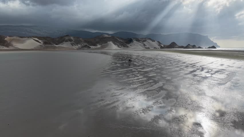 Wide coastal landscape with wet reflective beach, dark mountains and sand dunes under dramatic cloudy sky with sun rays, calm moody atmosphere, lots of copy space.