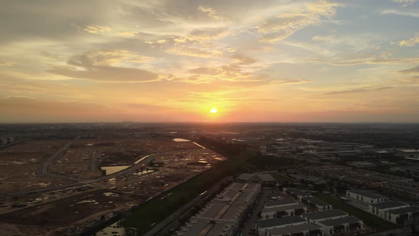 Aerial drone view of a beautiful golden sunset over an urban residential development area in Malaysia, featuring warm evening light, dramatic clouds, reflective water surfaces, and peaceful suburban cityscape scenery during dusk.