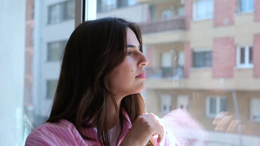 Thoughtful young woman with long brown hair looking out the window, lost in her thoughts and contemplating life. A moment of introspection and reflection as she observes the city outside her home