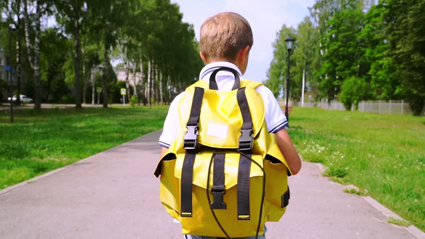 Boy walks down park path with bright yellow backpack. Sunlight filters through green trees beside him. He wears white shirt and moves calmly, purposefully