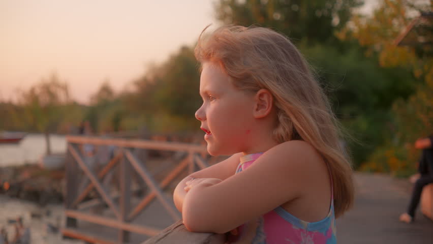 Child stands on a wooden boardwalk near water while people relax in the background during sunset hours