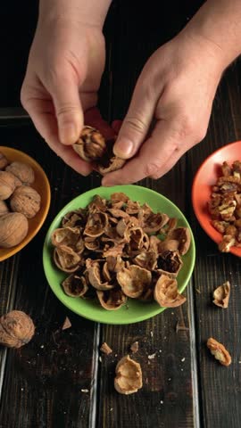 Hands are seen cracking walnut shells above a green bowl filled with empty shells, with whole walnuts and a bowl of walnut pieces visible in the surrounding area