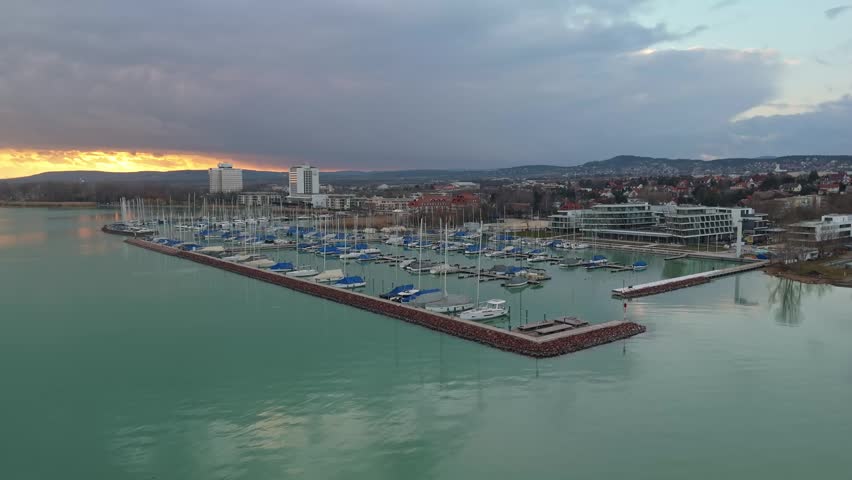 In Balatonfüred on Lake Balaton, a low-level orbital aerial view captures moored sailboats along the marina beside the urban waterfront with calm water, distant hills, and an overcast sky.