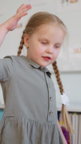 Young blonde girl with braided pigtails in a gray dress raising her hand and looking aside in a bright classroom near a window