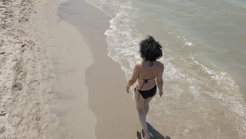 Aerial view of sexy woman in bikini walking along a sandy beach by the water.