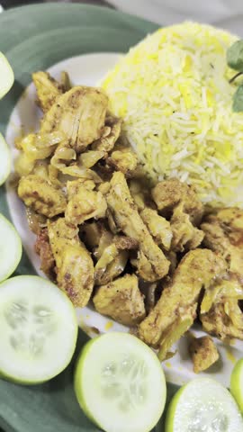 Overhead view of a hand carefully placing fresh cucumber slices as a garnish around a serving of domed yellow basmati rice and stir-fried spiced chicken pieces on a round green ceramic plate..Arranging Cucumber Slices on Chicken and Rice Plate
