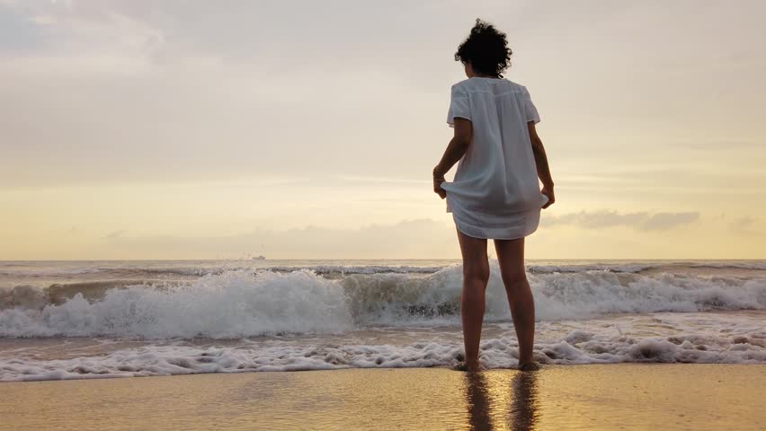 Woman in a white dress stands in the surf as waves splash around her legs