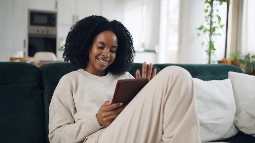 Relaxed young woman with afro hair smiling and scrolling on a digital tablet, sitting comfortably on a sofa in her modern living room