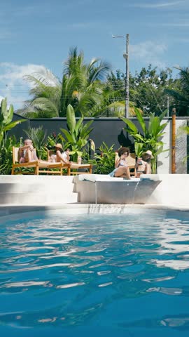 Four diverse women in swimsuits sunbathing and relaxing on lounge chairs by a luxurious swimming pool with a small waterfall on a sunny day. Vertical