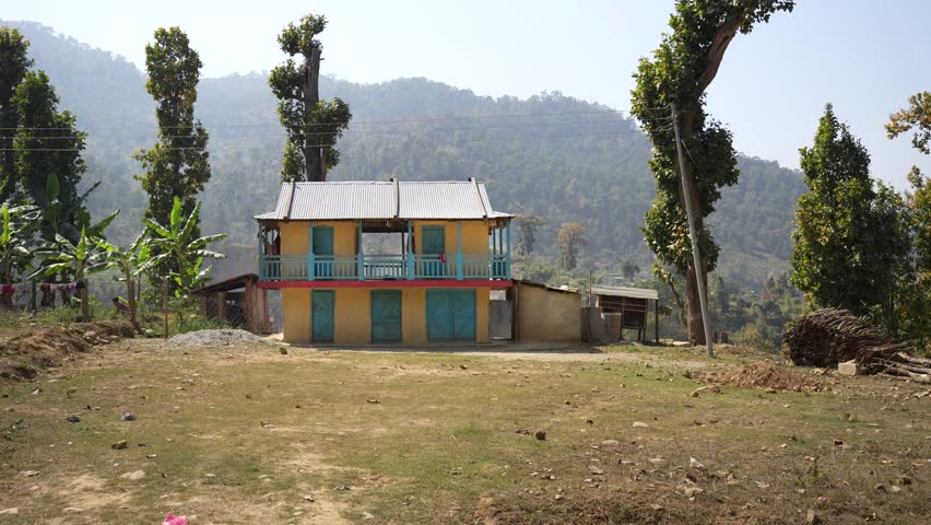 View of a traditional two-story house with yellow and blue walls surrounded by lush trees and rolling hills in a rural landscape, Nepal.
