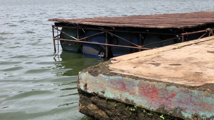 Rusty floating dock near weathered concrete pier on lake. Old wooden platform with blue barrels beside calm water. Aged harbor edge showing moss stains under sunlit waves