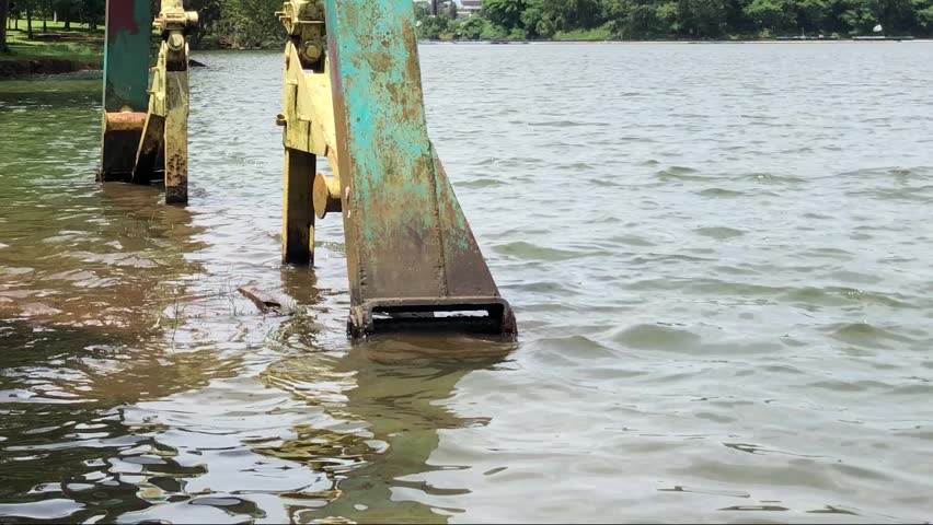 Weathered metal supports rising from lake water. Rusty pier pillars beside calm urban shoreline. Aged steel structure on tropical park waterfront