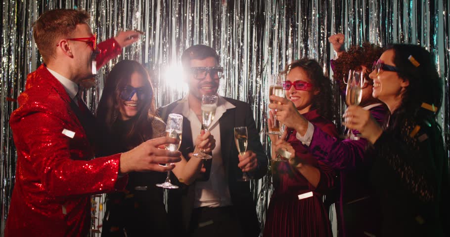 Group of friends celebrating together at disco party, wearing colorful retro outfits and funky glasses. Joyful people cheering, holding glasses of champagne and having fun against silver background.