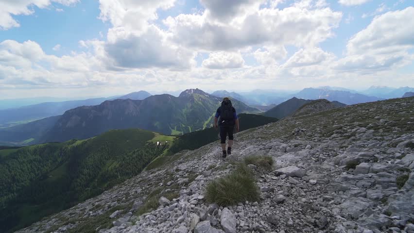 Wide view of an open green mountain plateau and trail with a dramatic cloudy sky above the distant mountain tops.