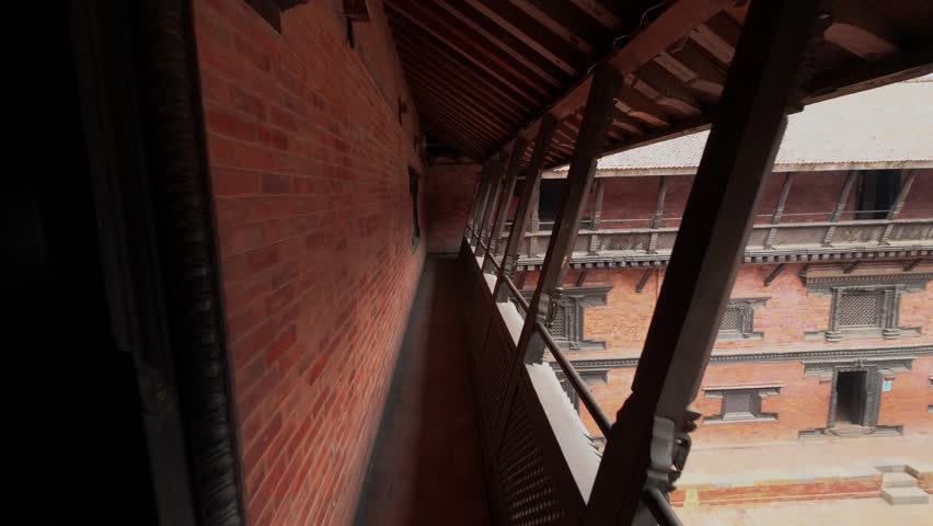 View of a narrow corridor with red brick walls and intricate dark wood carvings overlooking a courtyard in a historic building Nepal.