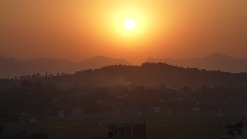 View of the sun setting over silhouetted mountain ridges and a hazy valley with small village buildings in Nepal.