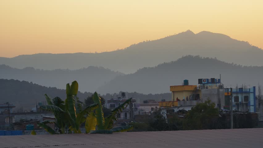 View of hazy mountain peaks and urban rooftops under a warm orange sky at sunset in Nepal.