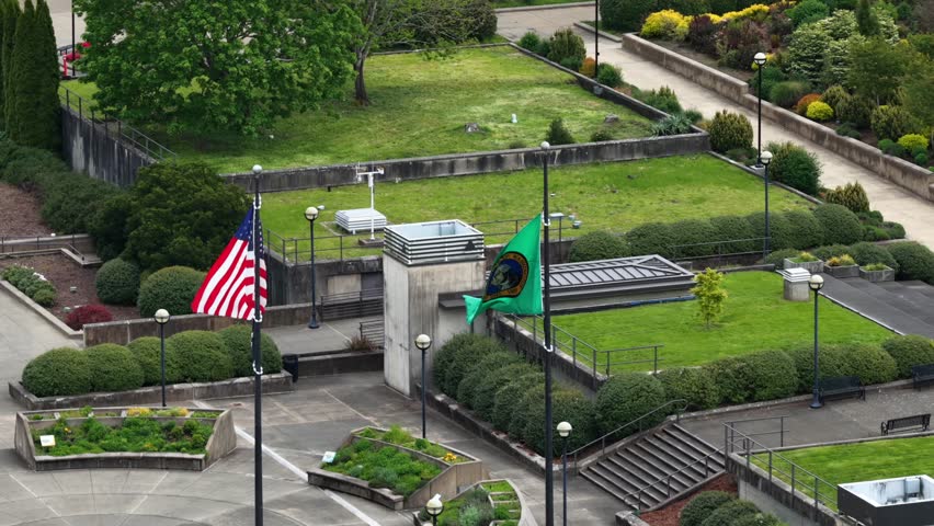 United States and Washington State flags waving on flagpoles at State Capitol campus in Olympia surrounded by green lawns and landscaped gardens during spring in United States. Aerial approaching.