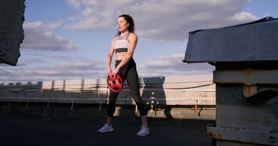 Sportive woman. Weighted plate lifting. Motivated athlete enjoying healthy activity training on roof holding red circular equipment.