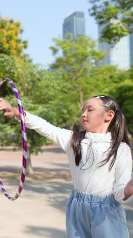A young girl in pigtails spins a purple hula hoop outdoors in a sunny park