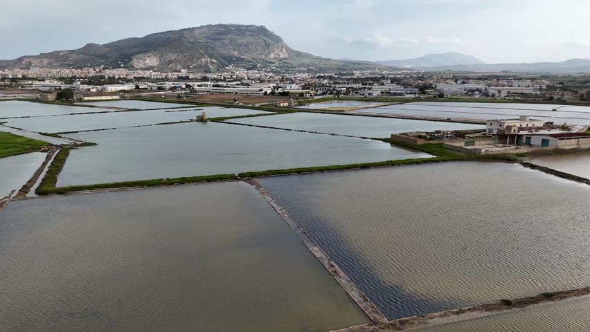 Drone view of Salina Galia Canino salt pans near Trapani, Sicily, with flooded basins, roads, city buildings, and mountain landscape.