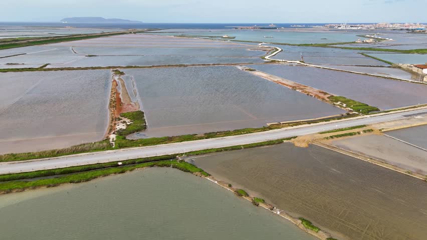 Drone panorama of Salina Galia Canino salt pans near Trapani, Sicily, with mountain skyline, cloudy sky, city edge, and coastal basins.