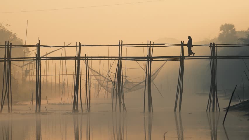 View of a person standing on a traditional bamboo bridge over a misty river with fishing nets at sunrise in Bogura, Rajshahi Division, Bangladesh.