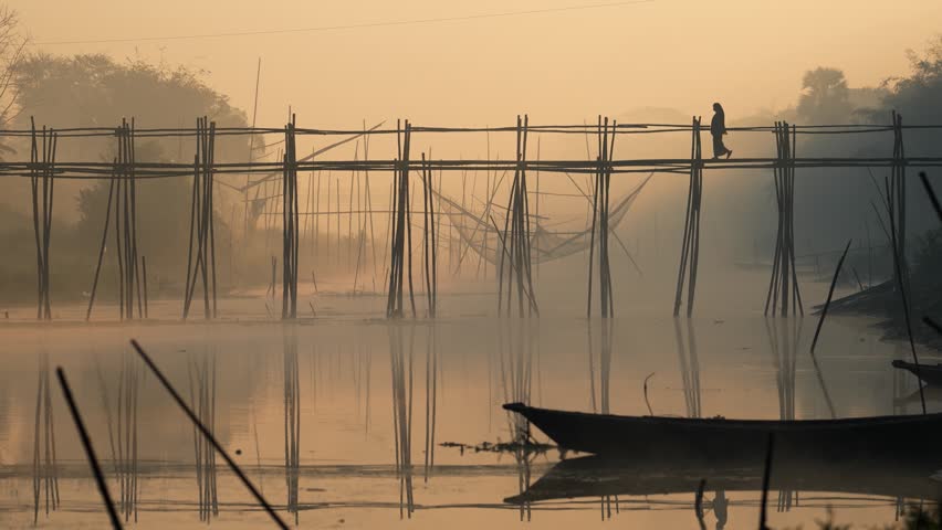 View of a person standing on a bamboo bridge over a misty river with fishing nets and a boat at dawn in Bogura, Rajshahi Division, Bangladesh.