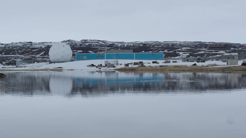 Ground-level camera shot of a large white radome dome and blue industrial building perfectly reflected in a calm pool of water during spring thaw in Iqaluit.