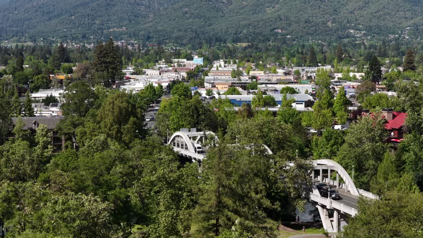 Grants Pass Oregon downtown with historic arch bridge over Rogue River, lush green tree canopy, low-rise commercial district, residential neighborhoods and forested hills under sunny sky. Aerial rise