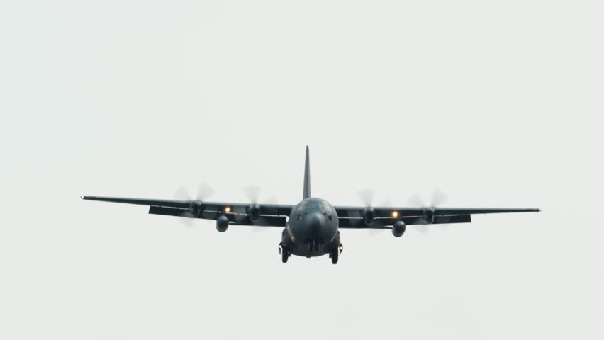 Military C-130 Hercules cargo aircraft in flight carrying equipment and personnel during defense operation. Tactical transport airplane against gray overcast sky.