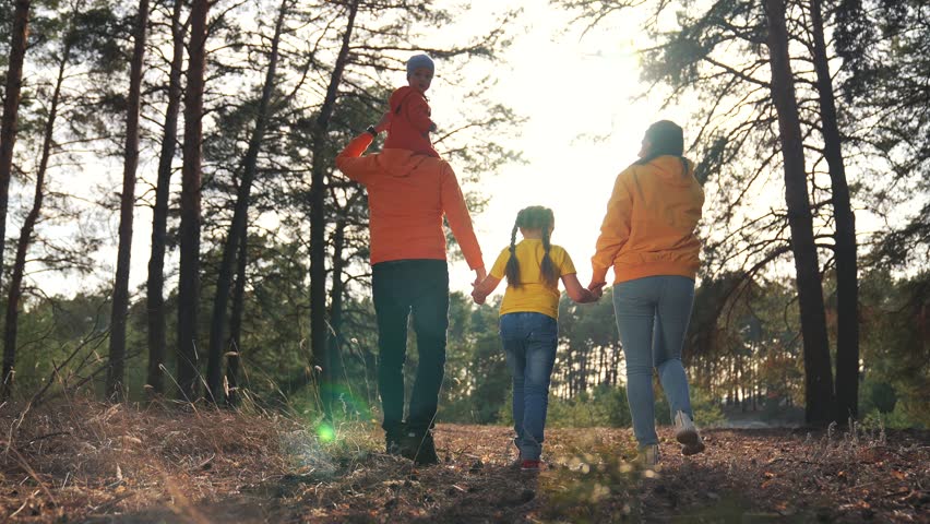 Family takes walk through nature forest. Father carries young child on shoulders. Mother holds daughter hand during walk. Happy family explores outdoor nature. Parents feel calm in pine forest.