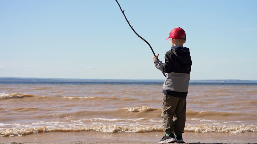 Young boy in a red cap standing on a sandy shore and playing with a big stick, pretending to fish as waves from the murky water splash near his feet on a bright, sunny day by the sea