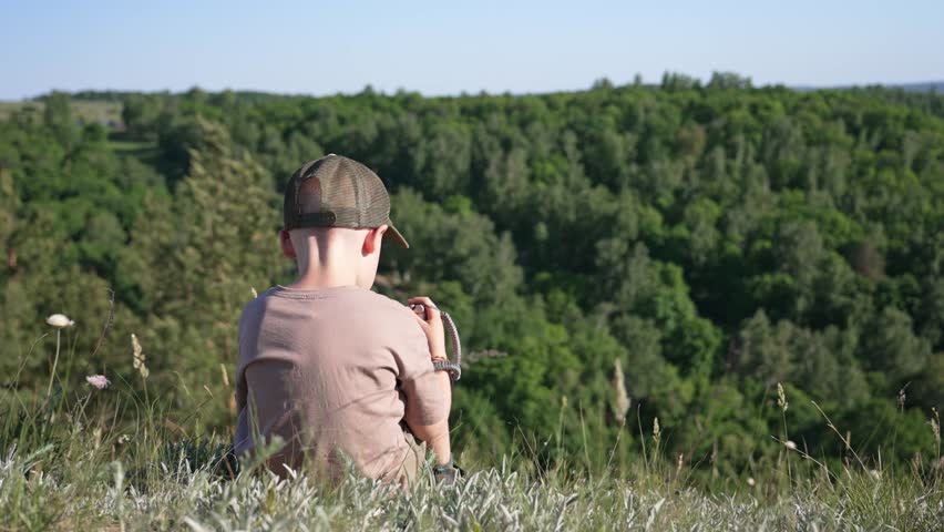 Lonely young boy wearing a cap sitting on the edge of a grassy hill, peacefully looking out over a vast green forest on a sunny day, representing a moment of quiet contemplation