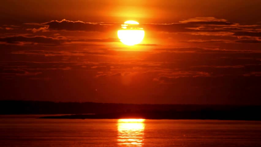 A bright golden sun sets over calm water, casting a vibrant orange glow and reflection across the horizon