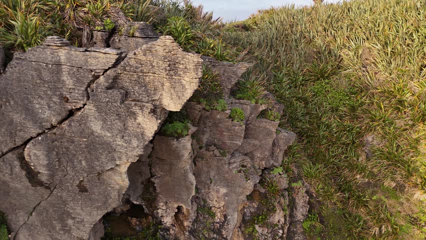 Drone panoramic shot glides from wide to close-up on Pancake Rocks at sunset in Punakaiki, New Zealand West Coast. Waves crash on detailed layered limestone formations under golden skies