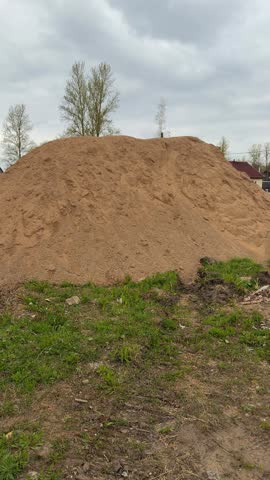 Large sand pile on construction site, spring day with overcast sky, grassy foreground and scattered debris, distant houses.