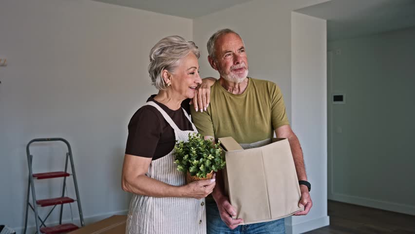 Happy elderly couple standing in their new empty apartment, holding a cardboard box and a plant, planning their future home together